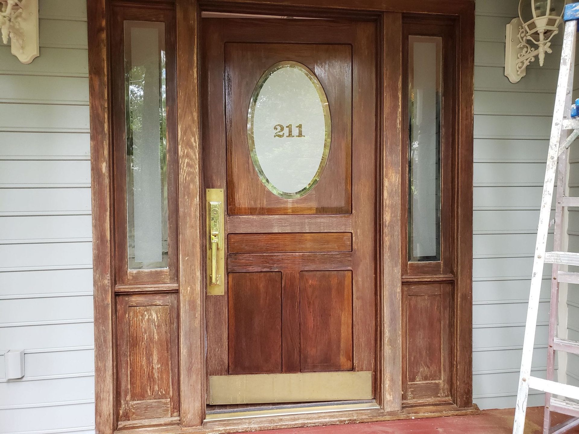 Wooden front door with side windows and address number 211, showing visible wear and tear, next to a ladder.