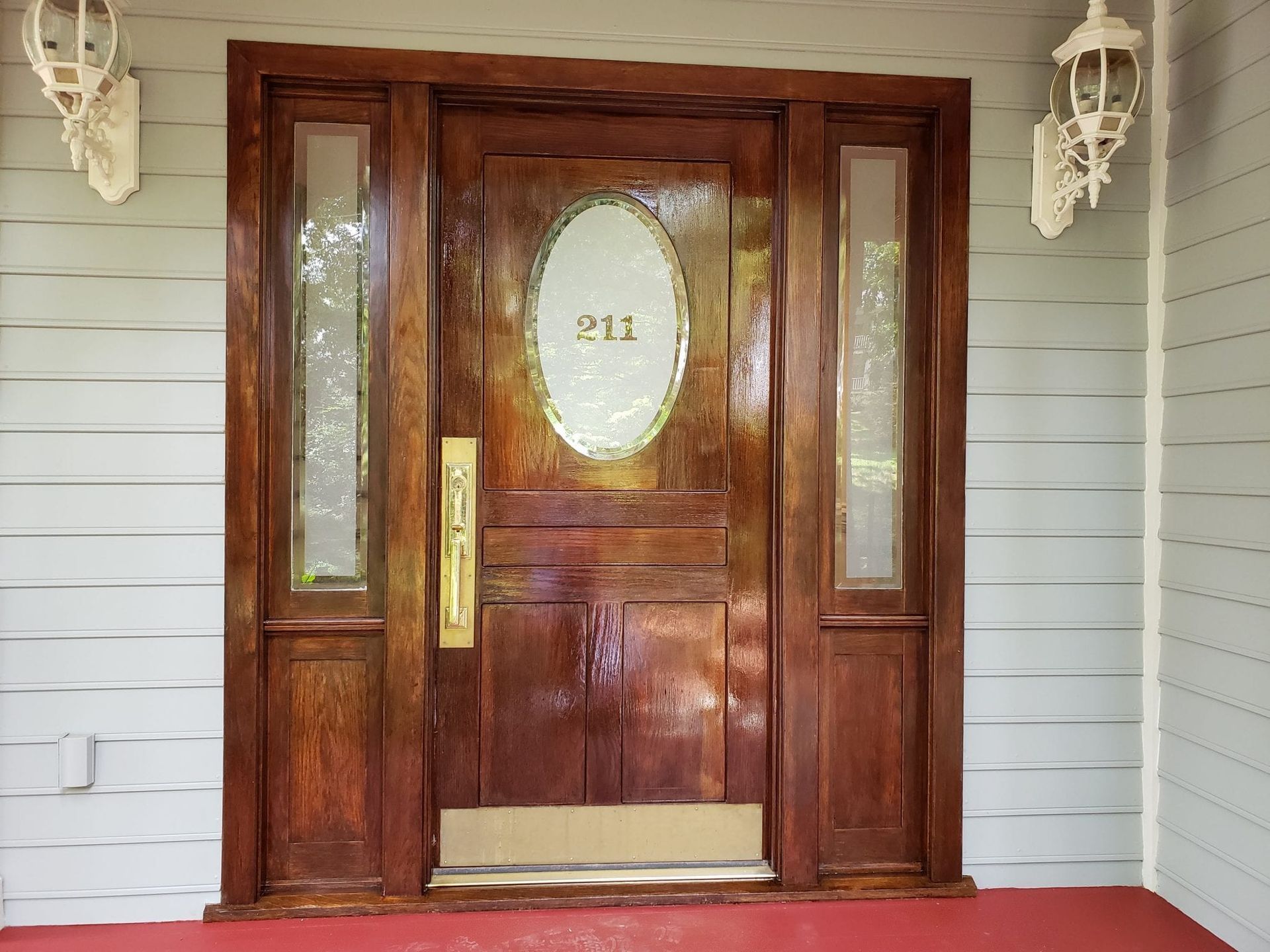 Wooden front door with sidelights, oval window, and house number 211, flanked by wall-mounted lanterns.