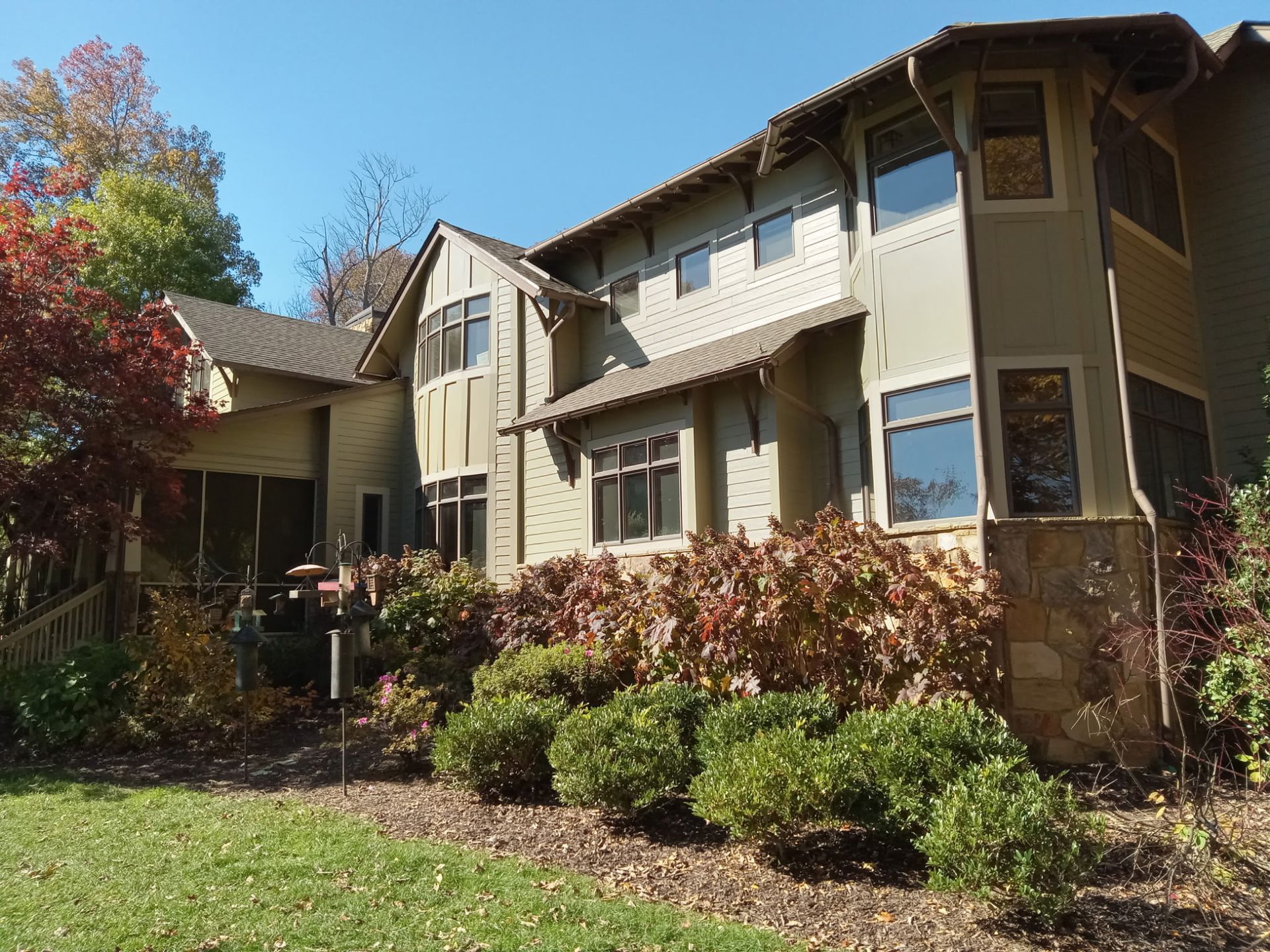 Two-story house with green siding, brown roof, and lush landscaping on a sunny day.