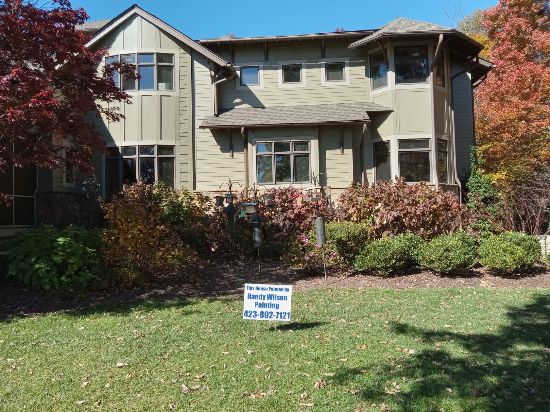 Two-story beige house with multiple windows, surrounded by landscaping and a for sale sign on the lawn.