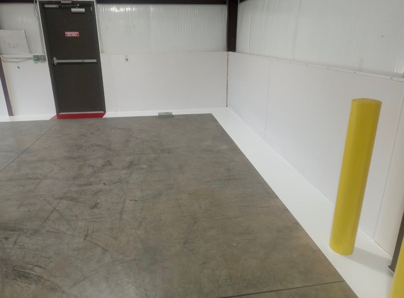 Empty warehouse interior with concrete floor, white walls, and a black door. Yellow bollards and safety strip on floor.