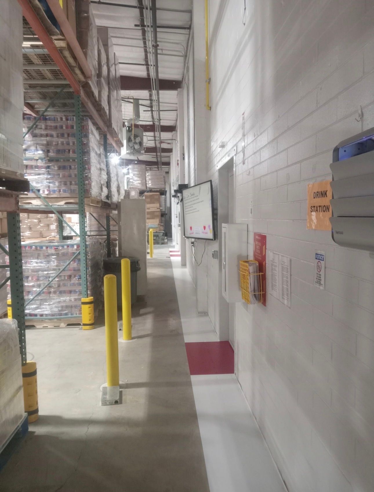 Warehouse aisle with shelving on the left, white wall on the right, and safety equipment.