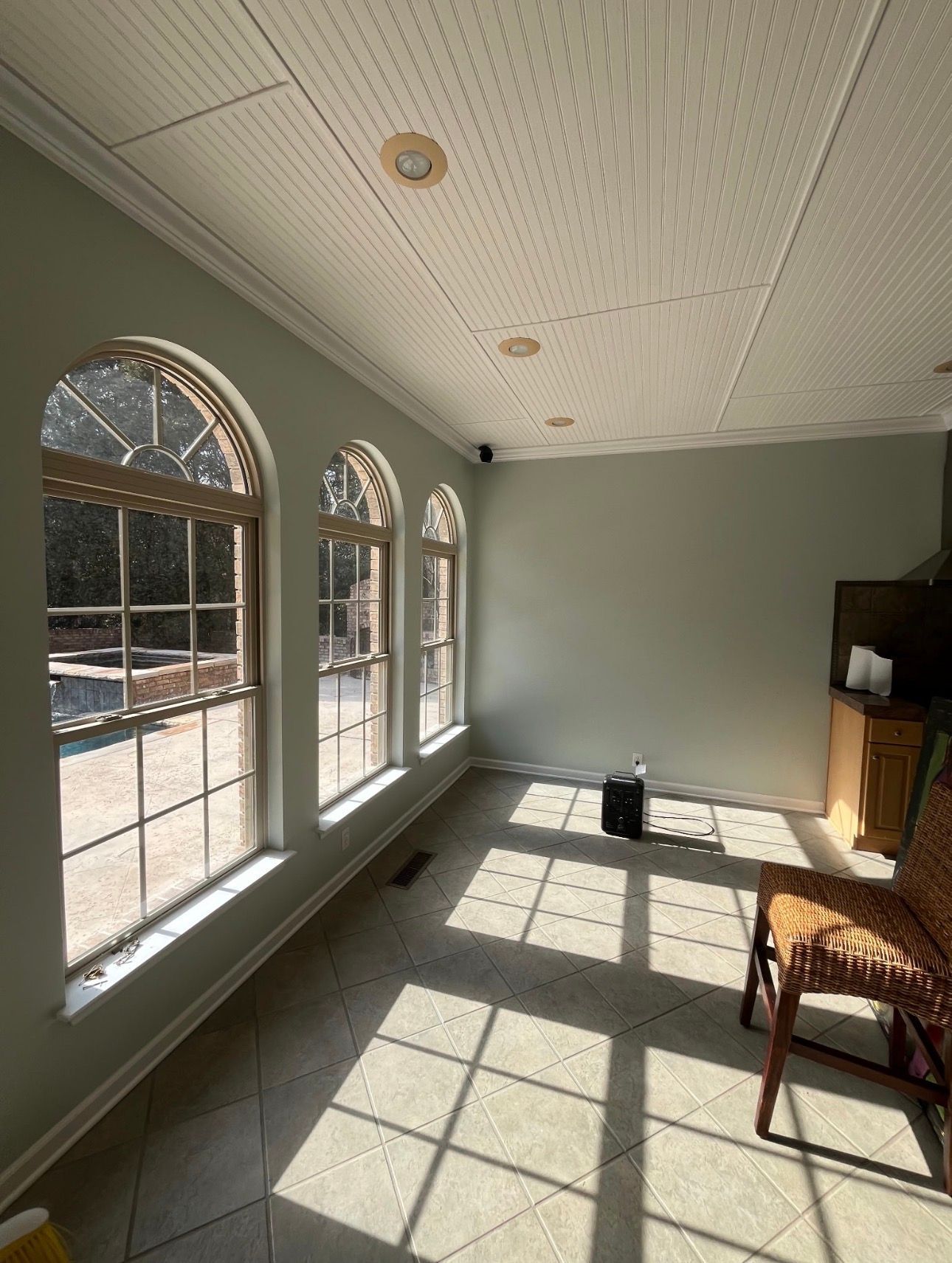 Sunlit room with arched windows, light green walls, patterned ceiling, tiled floor, and a wooden chair.