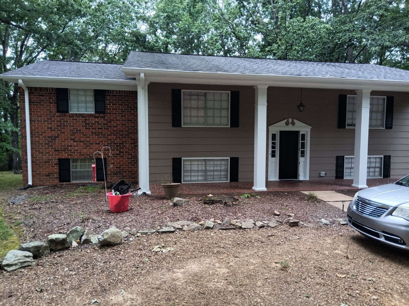 Two-story house with brown siding and brick, black shutters, white columns, and a gravel driveway.