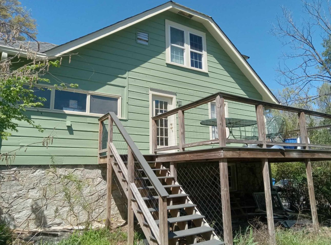Green house with wooden deck and stairs, door leading outside, stone foundation.