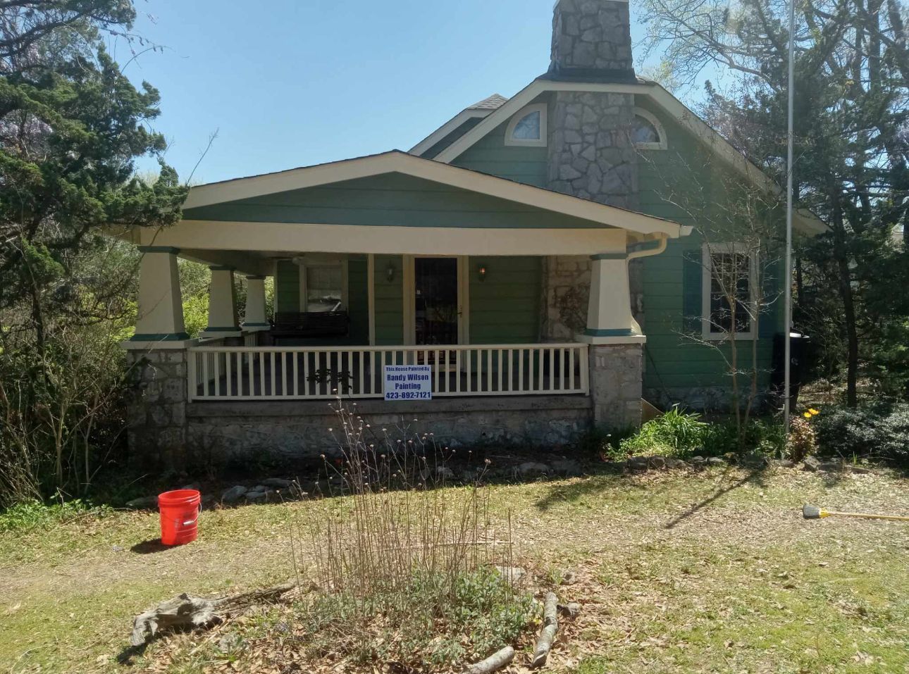 Green house with porch; stone chimney. A red bucket sits on the ground.