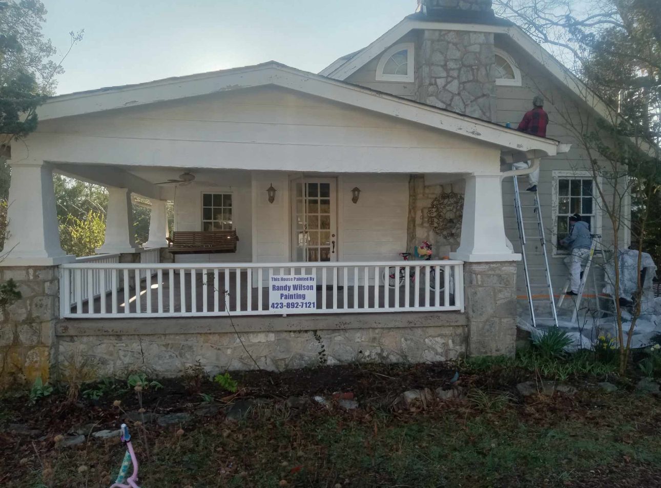 House being painted. White porch, stone chimney, ladder, person painting on roof.