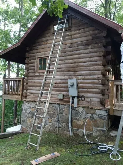 Log cabin with a ladder propped against it; stone foundation, electrical box, and deck visible.