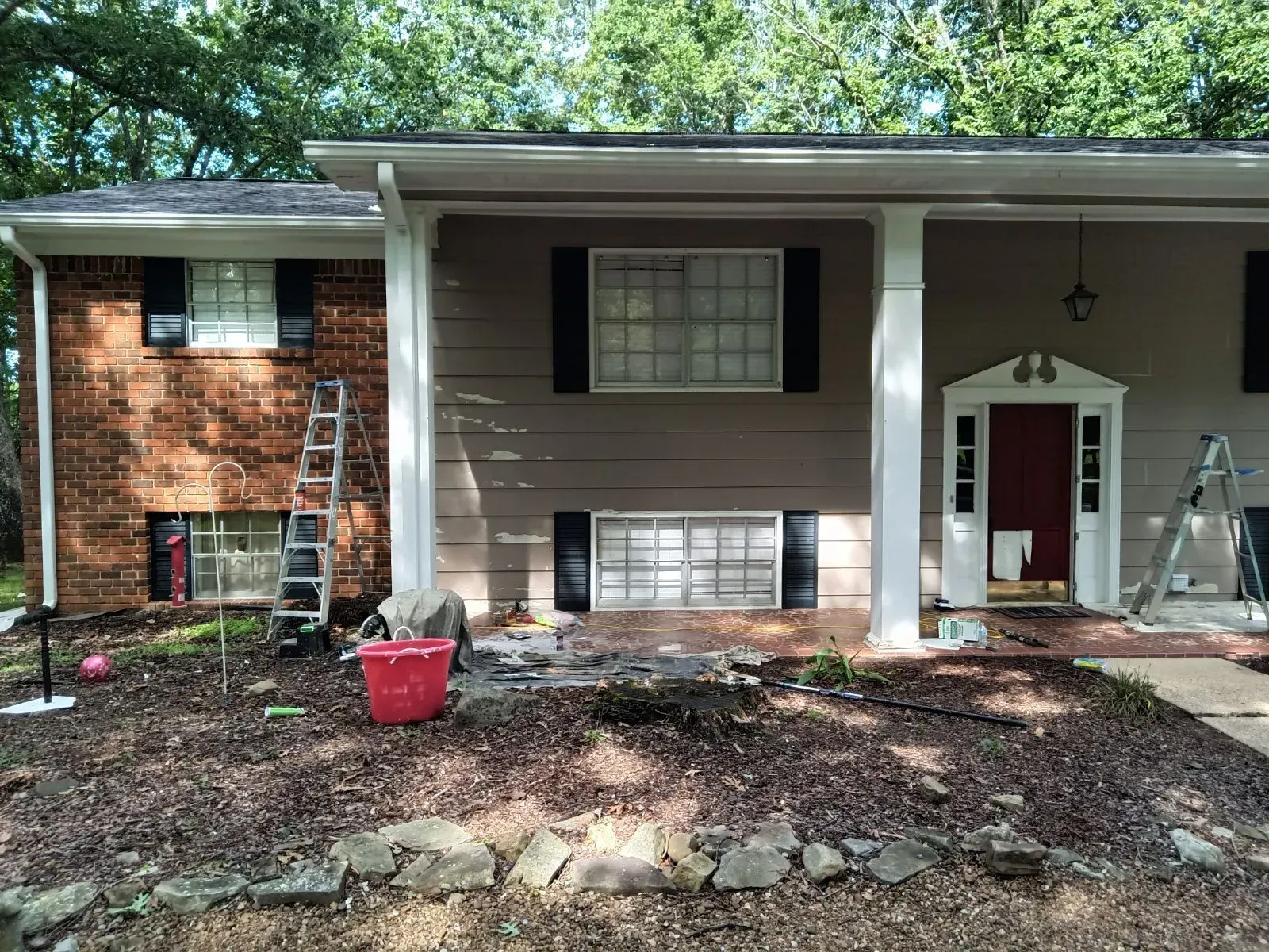 House exterior with brick and tan siding, columns, and a red door. Landscaping and ladder visible.