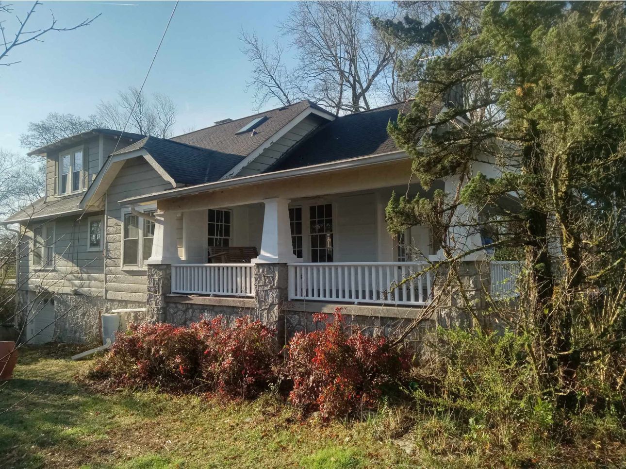 A two-story house with a porch and stone foundation, shrubs in front, and trees in the background.