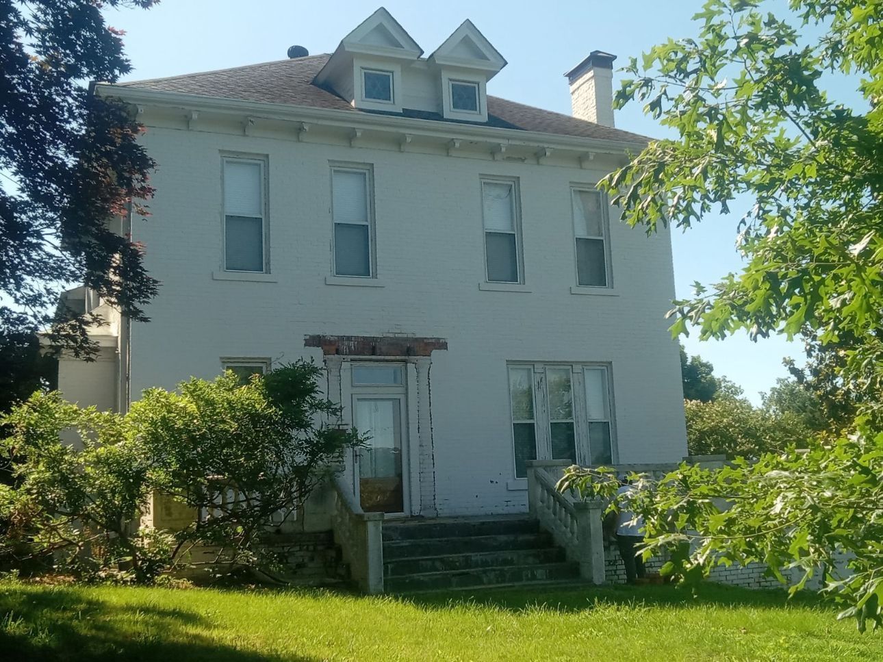 Two-story white house with peeling paint, dormers, and overgrown vegetation in front; steps lead to the entrance.