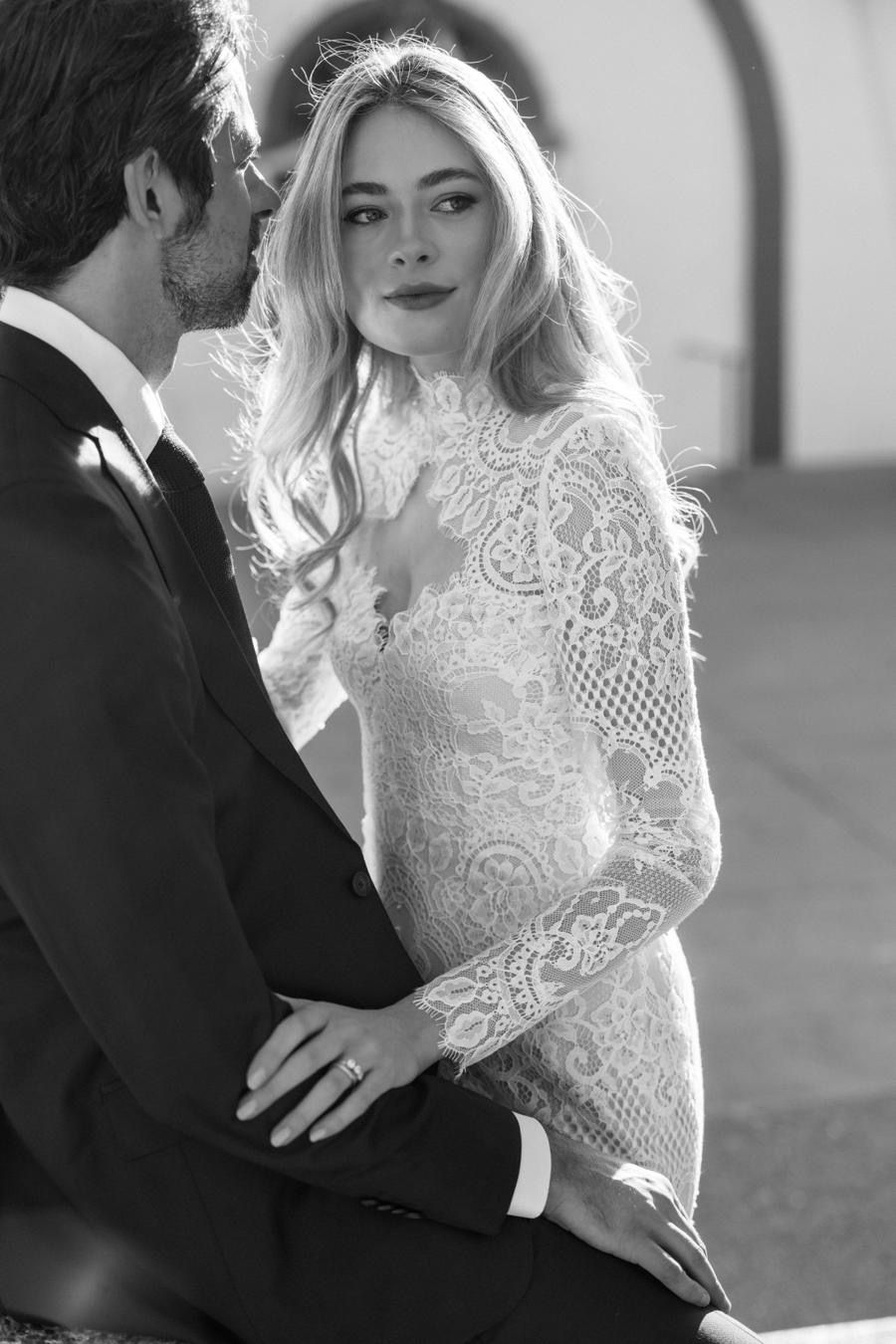 A bride and groom are kissing in a black and white photo . the bride is wearing a lace dress.