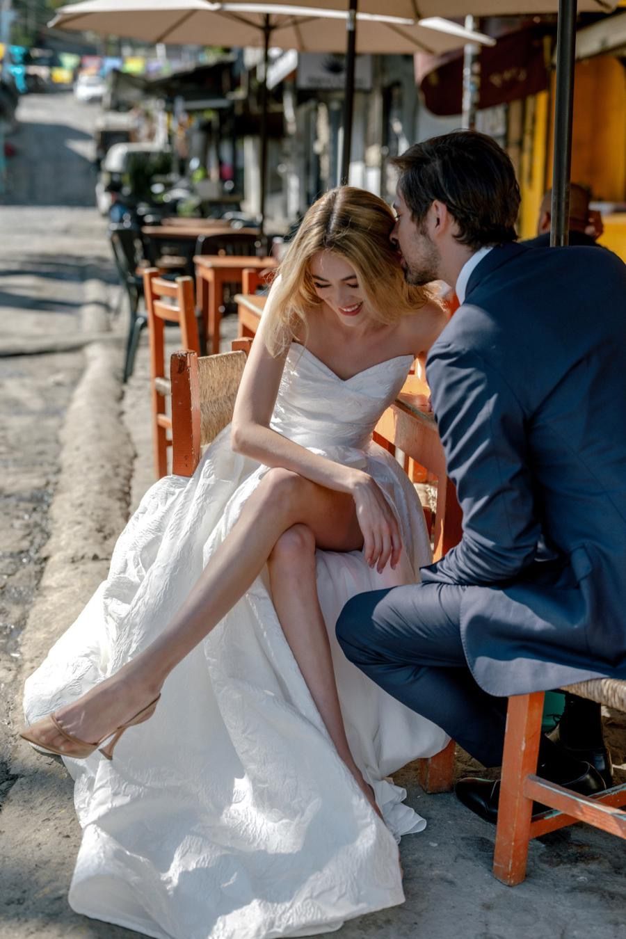 A bride and groom are sitting on a bench in front of a restaurant.