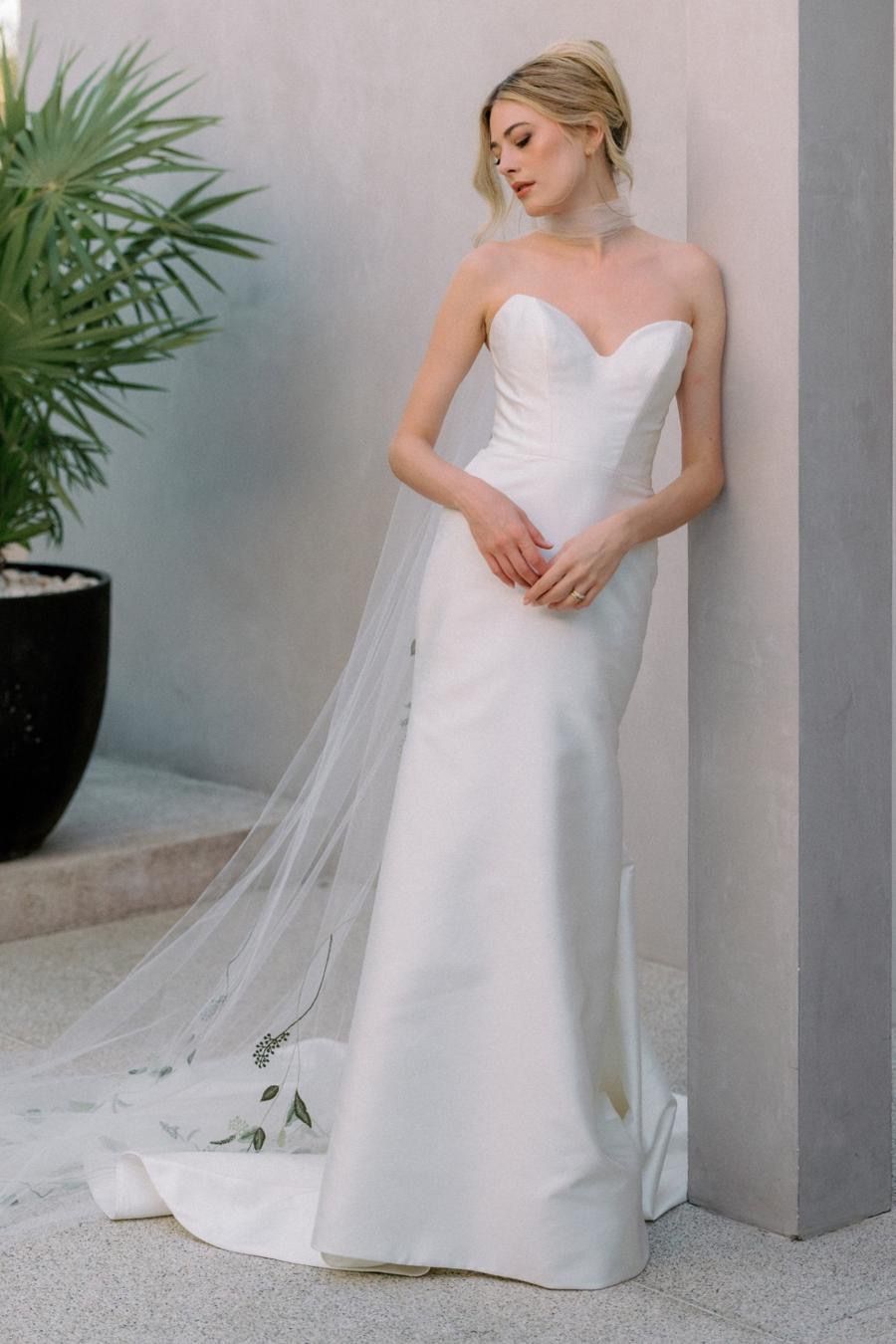 A woman in a white wedding dress is leaning against a wall.