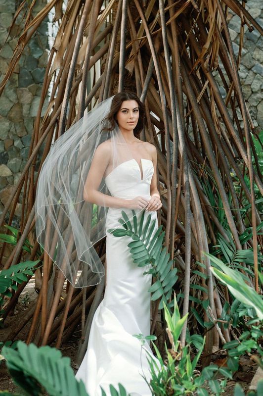 A woman in a wedding dress and veil is standing in front of a tree.