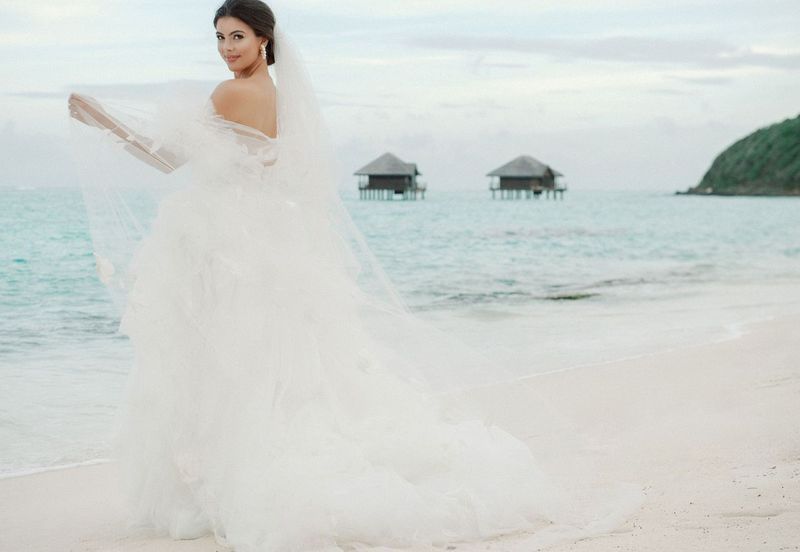 A bride in a wedding dress and veil is standing on a beach.