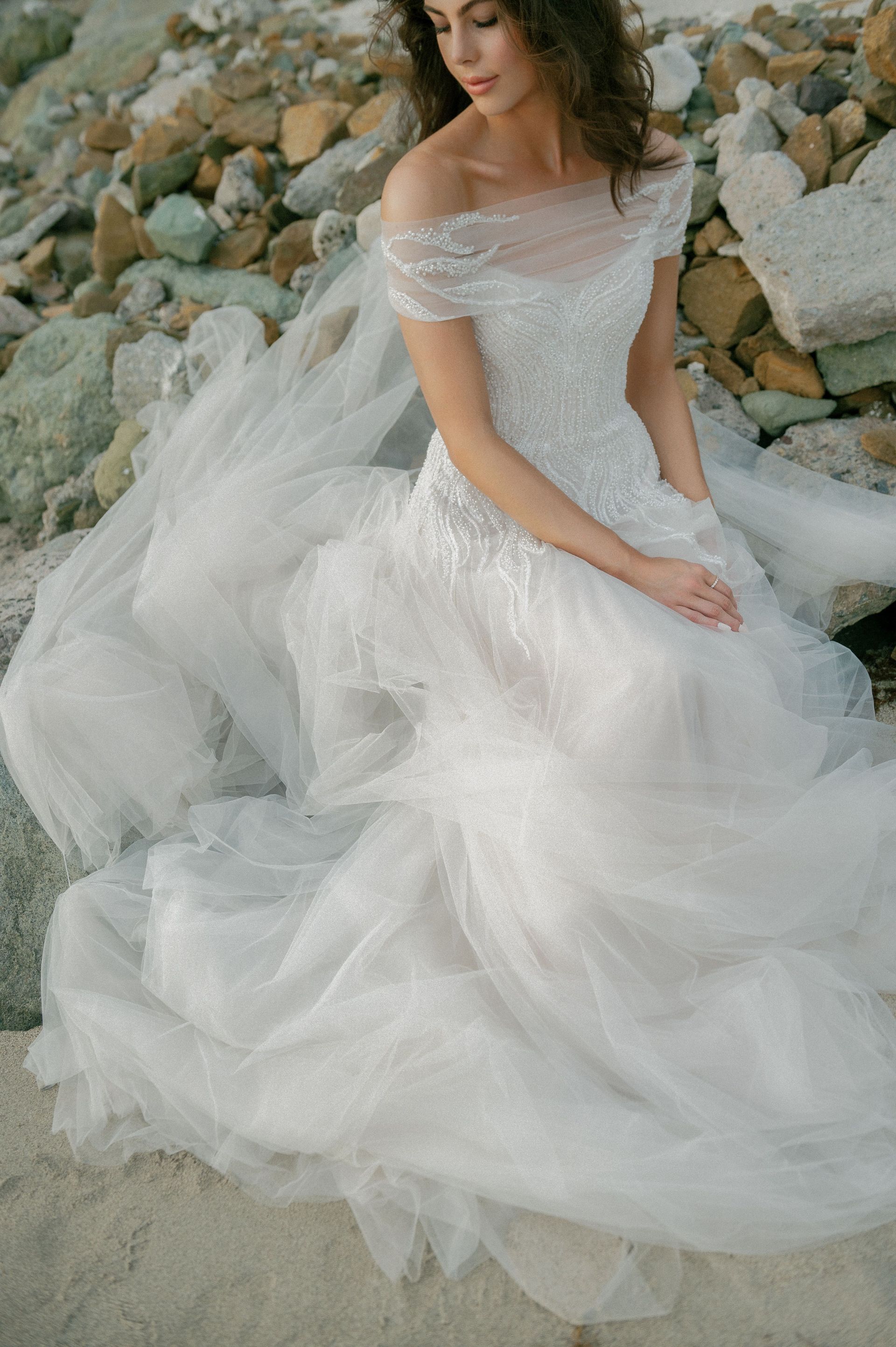 A woman in a wedding dress is sitting on a pile of rocks on the beach.