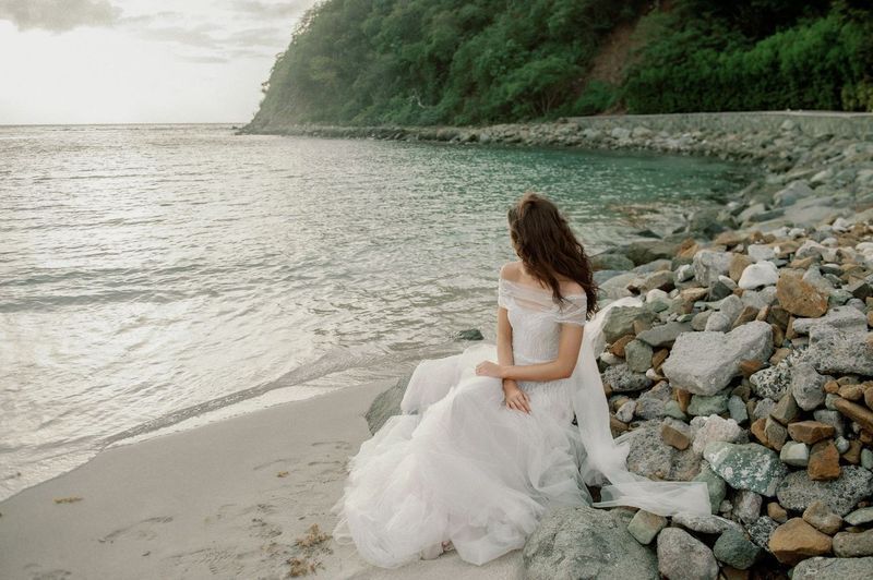 A woman in a wedding dress is sitting on a rocky beach looking at the ocean.
