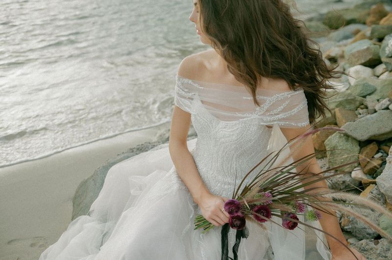 A woman in a wedding dress is sitting on the beach holding a bouquet of flowers.