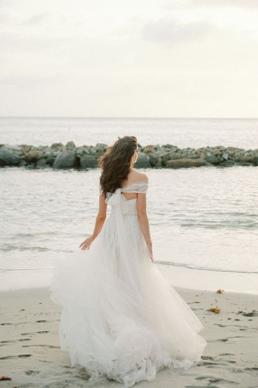A woman in a wedding dress is standing on a beach looking at the ocean.