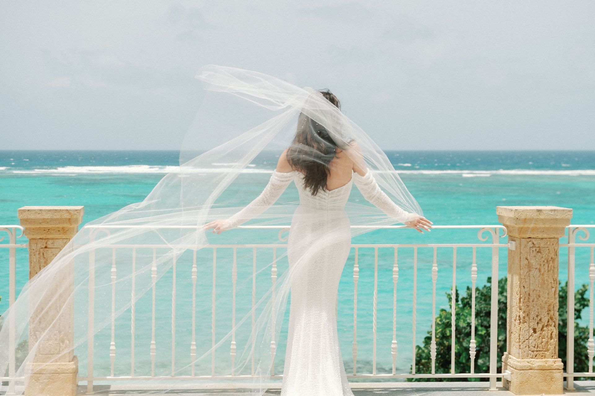 A bride in a wedding dress and veil is standing on a balcony overlooking the ocean.