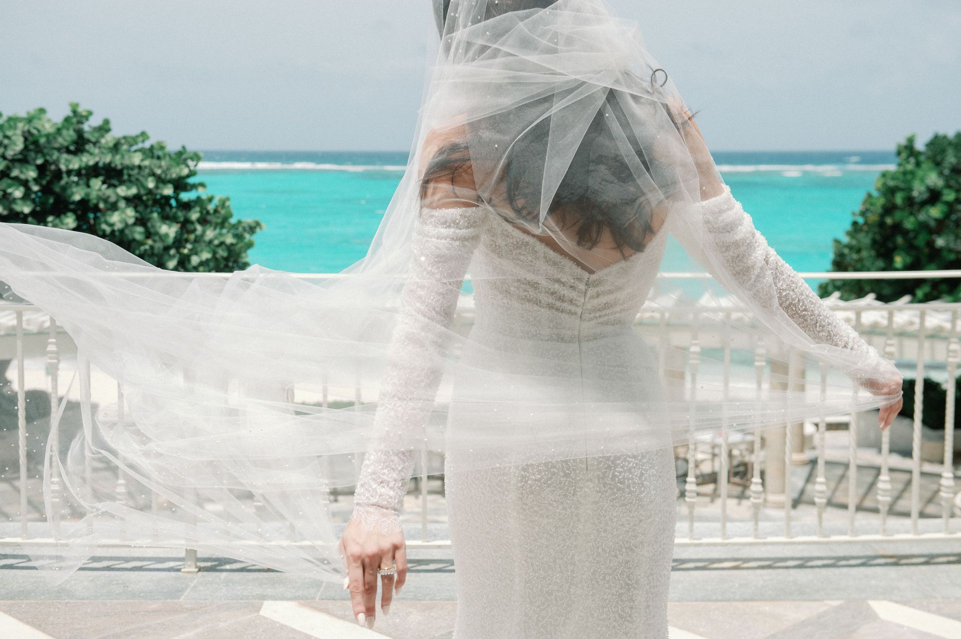 A bride in a white dress and veil is standing on a balcony overlooking the ocean.