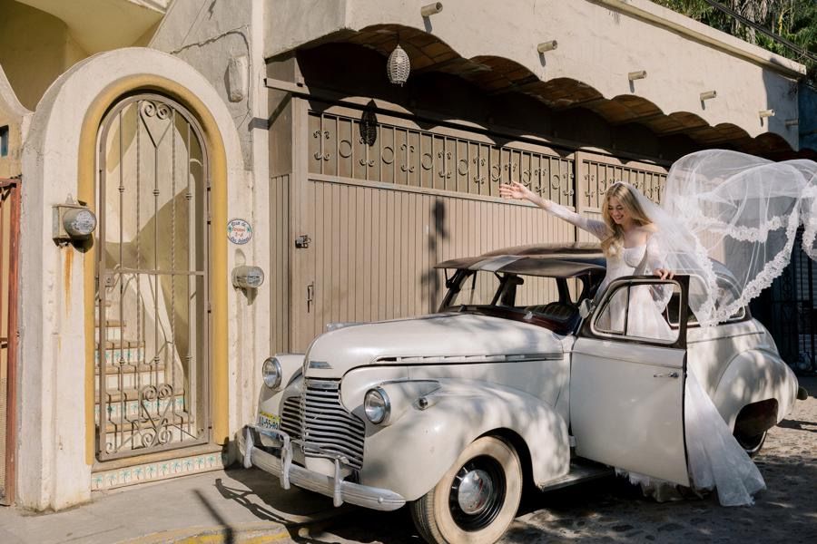 A bride in a wedding dress is standing next to an old white car.