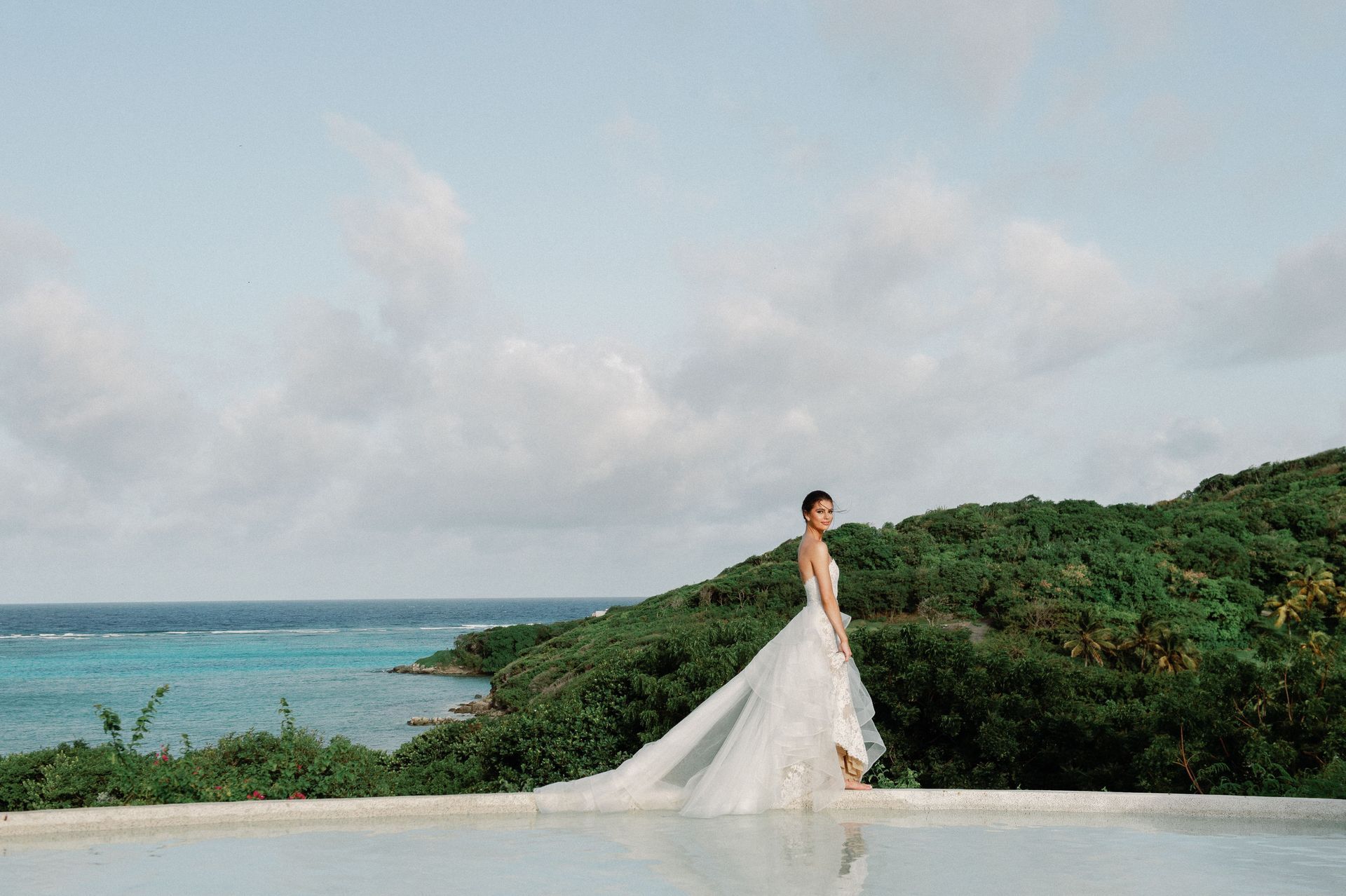 A bride in a wedding dress is standing next to a pool overlooking the ocean.