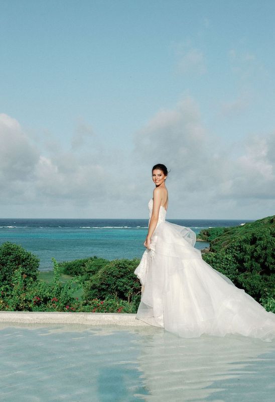 A bride in a wedding dress is standing next to a pool overlooking the ocean.