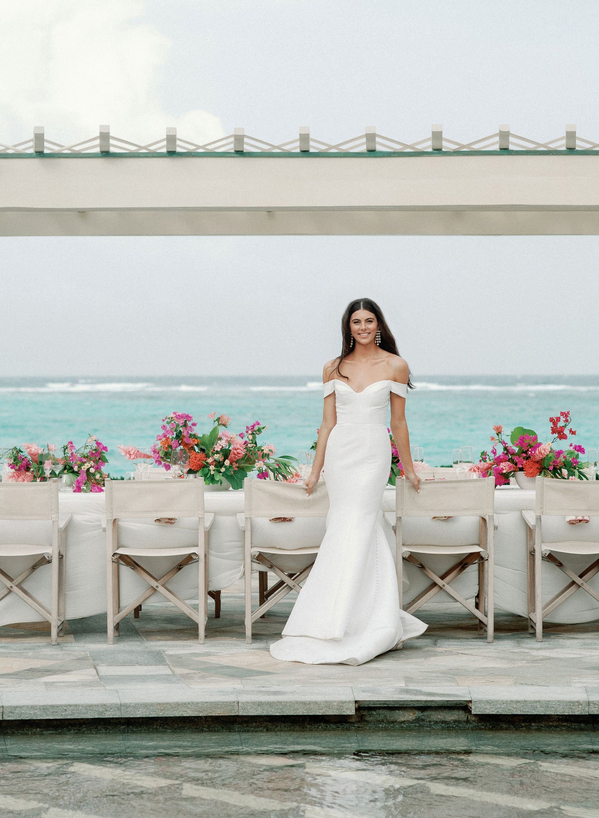 A bride in a white dress is standing in front of a table and chairs by the ocean.