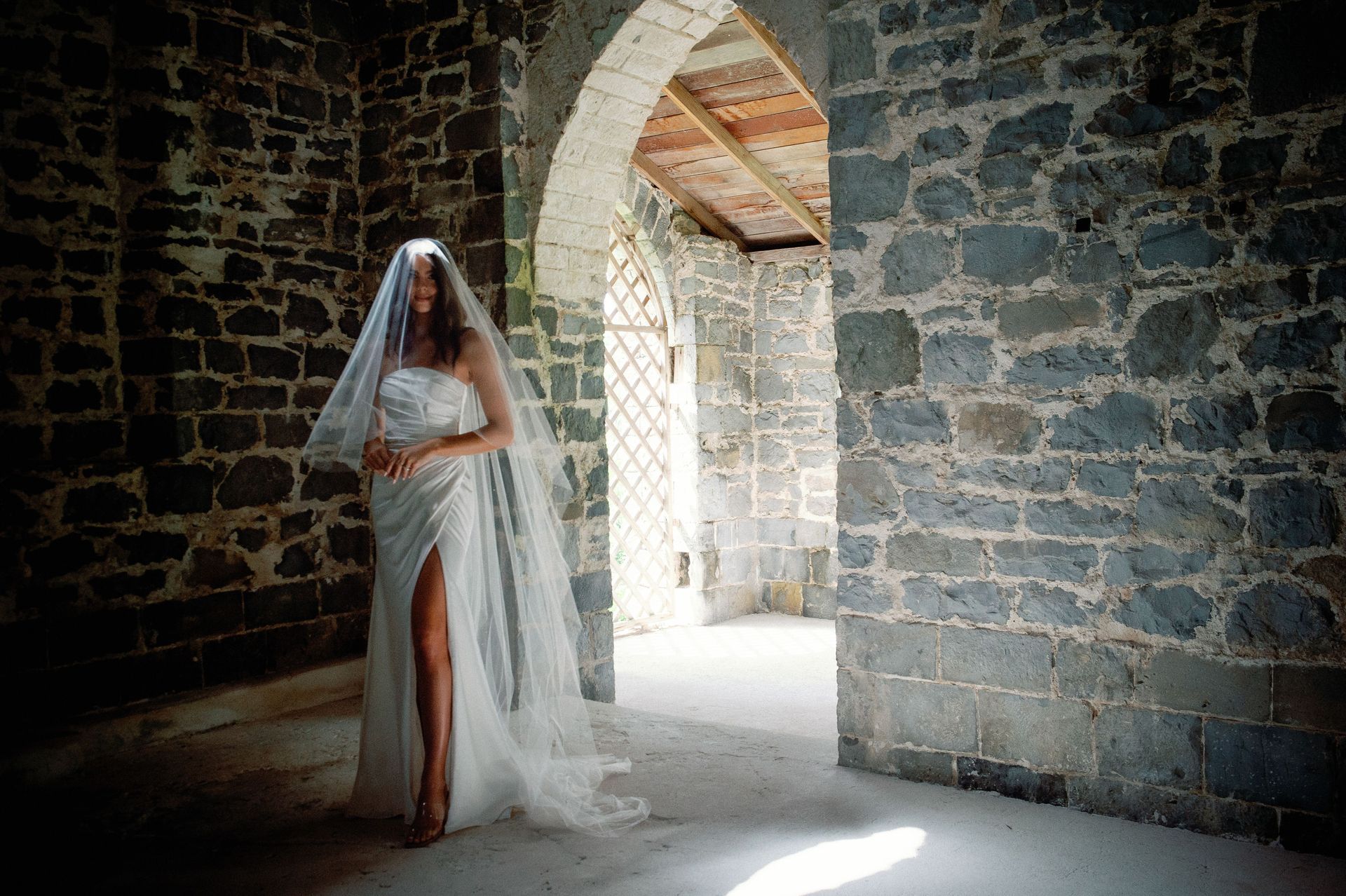A woman in a wedding dress and veil is standing in a stone building.