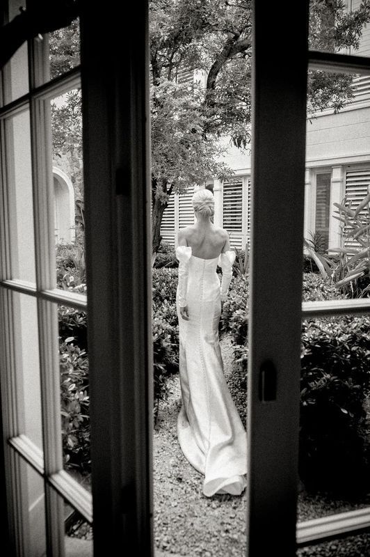 A black and white photo of a bride in a wedding dress standing in front of a window.