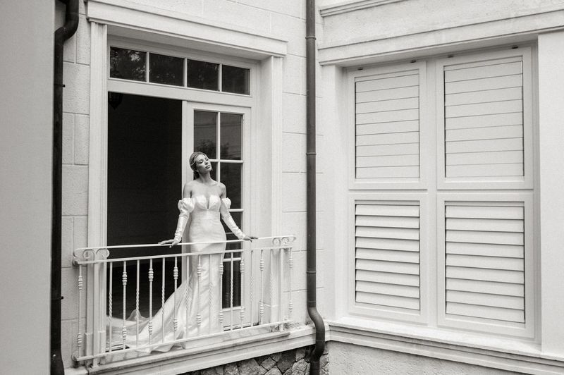 A black and white photo of a woman in a wedding dress standing on a balcony.