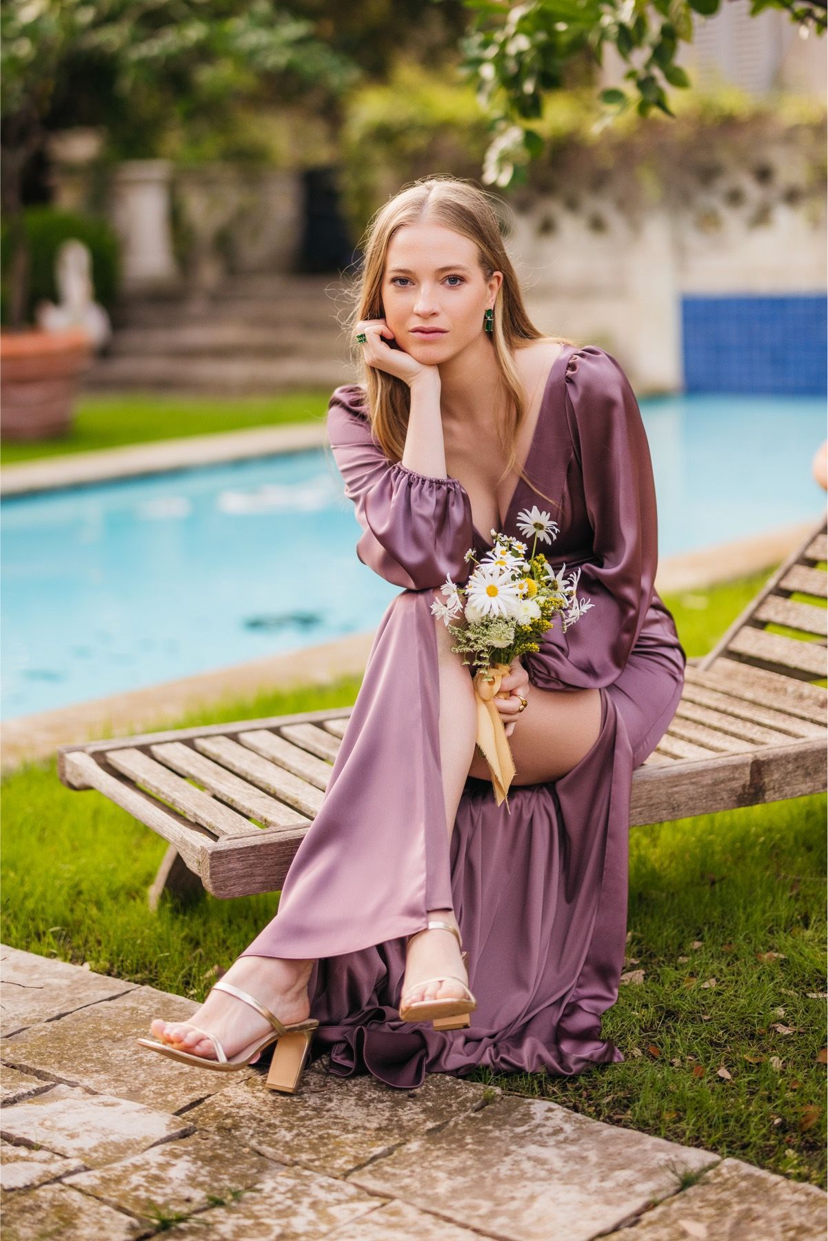 A woman in a purple dress is sitting on a chair next to a pool holding a bouquet of flowers.
