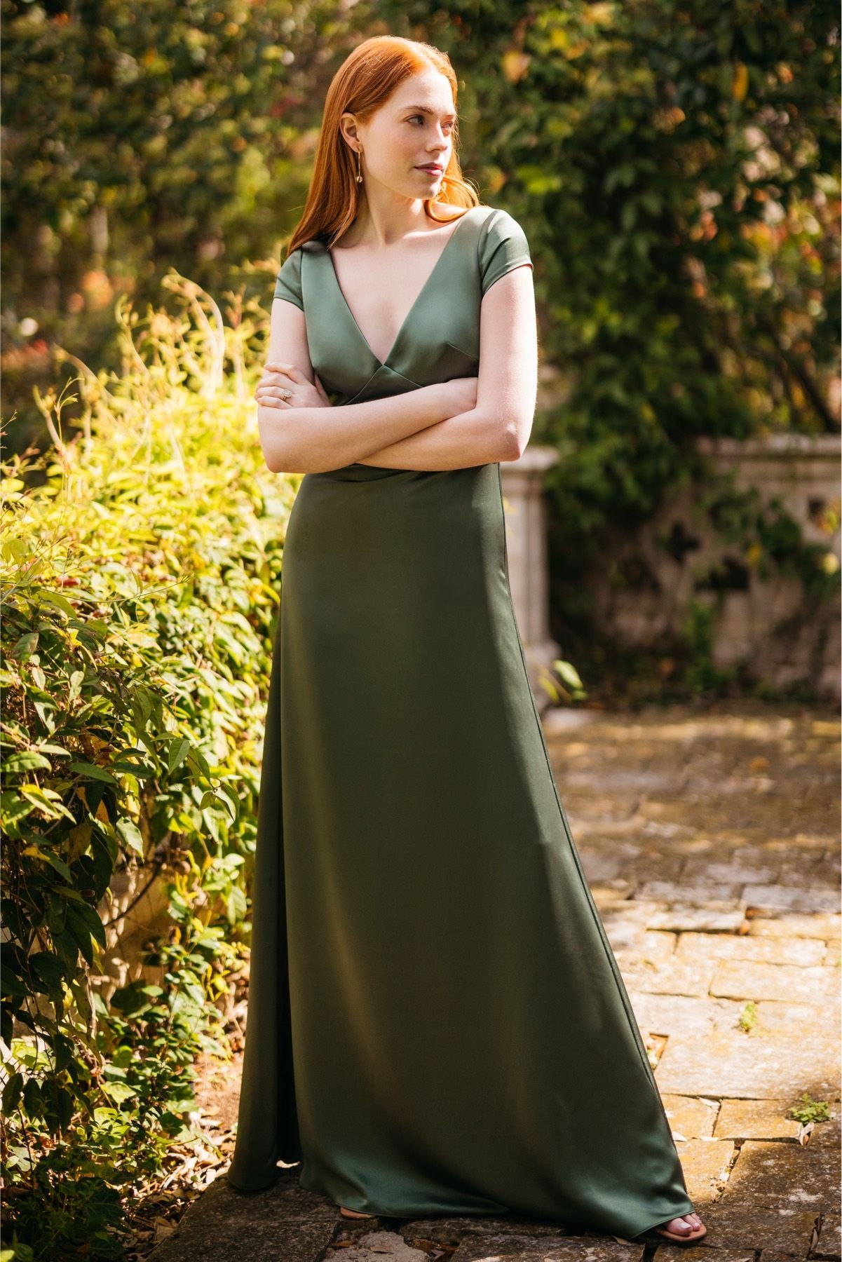 A woman in a green dress is standing in a garden with her arms crossed.
