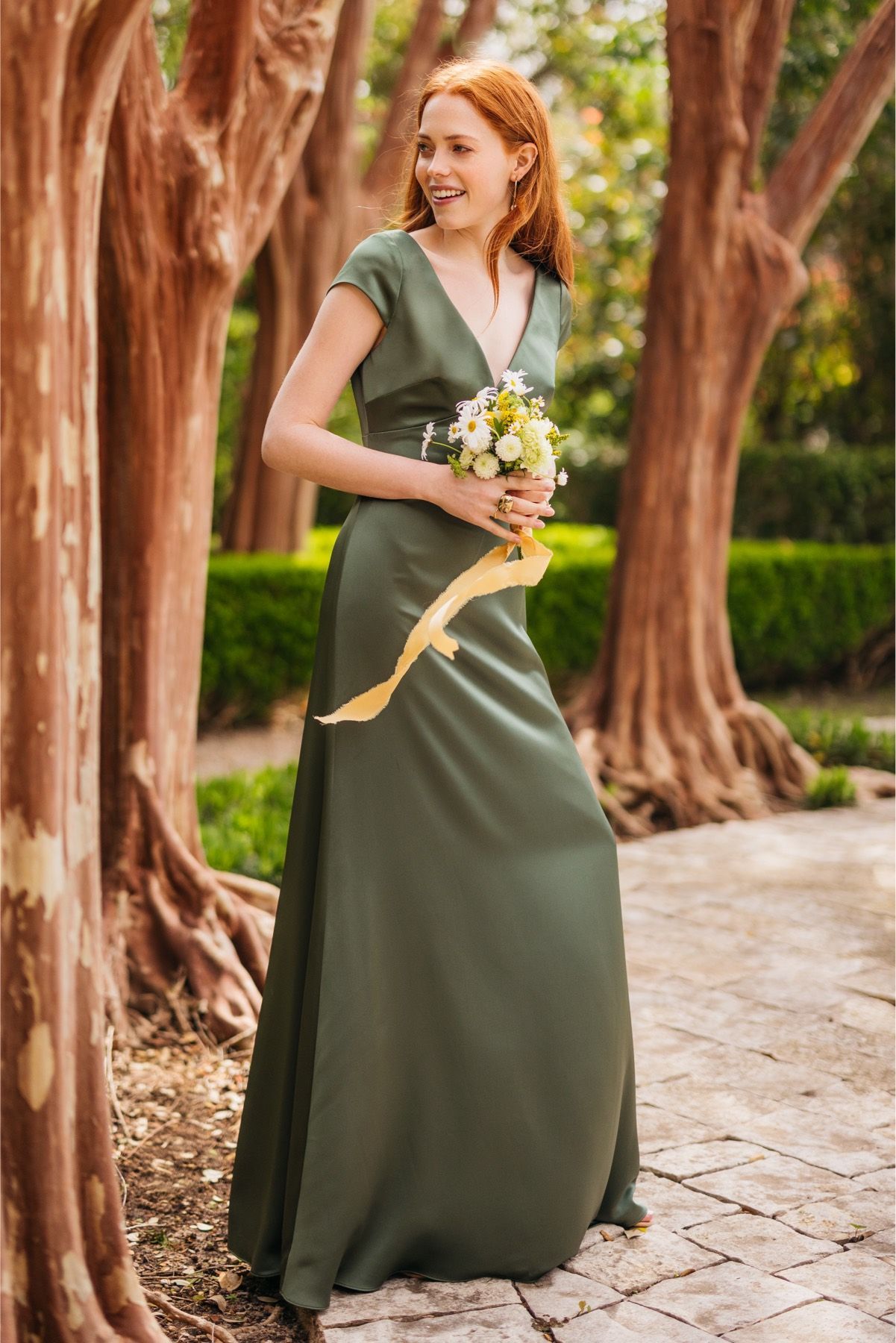 A woman in a green dress is standing next to a tree holding a bouquet of flowers.
