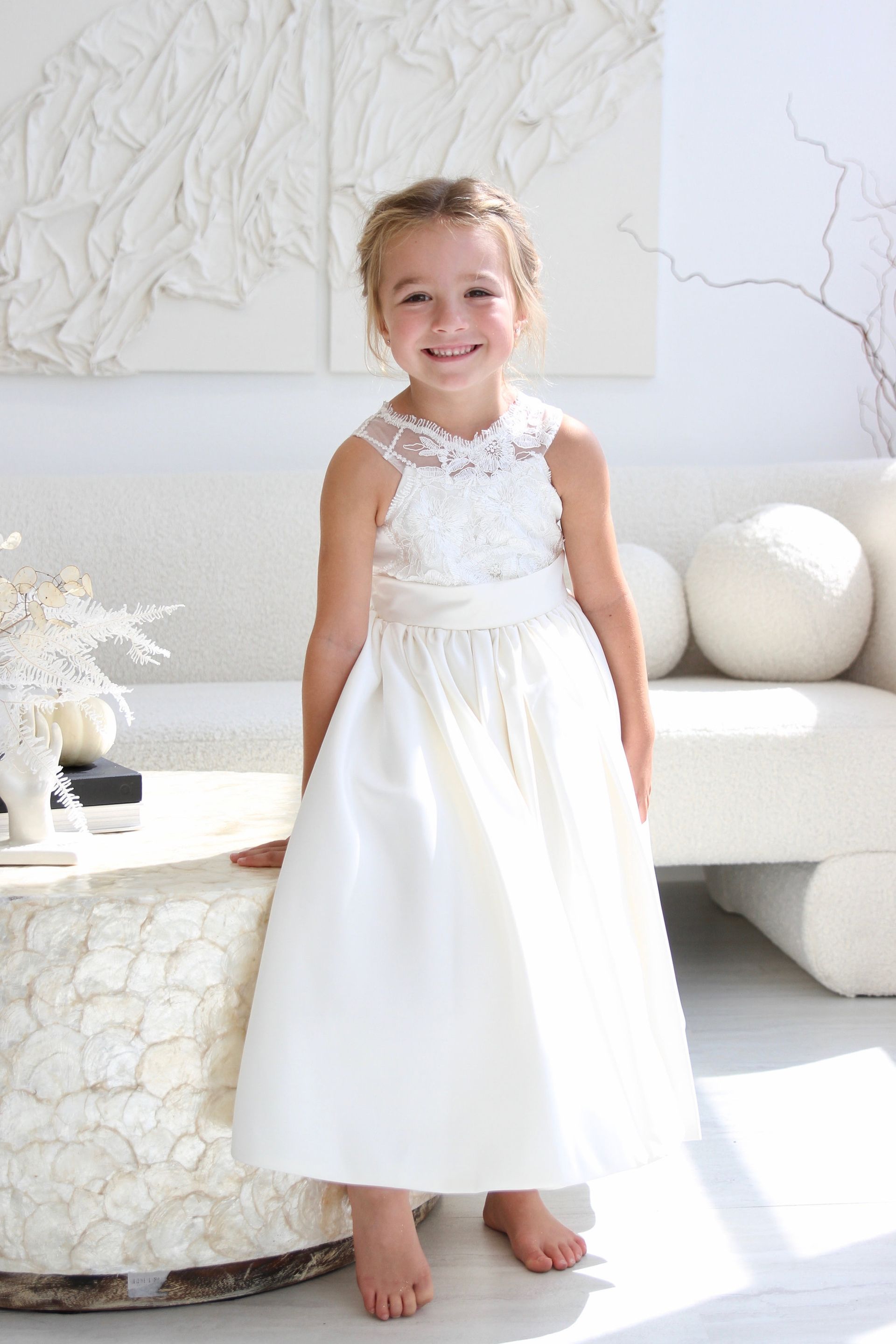 A little girl in a white dress is standing next to a table.