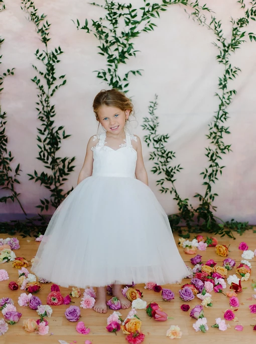 A little girl in a white dress is standing on a wooden floor surrounded by flowers.