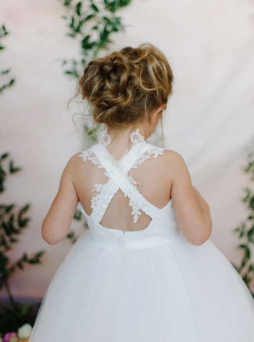 A little girl in a white flower girl dress is standing in front of a pink wall.