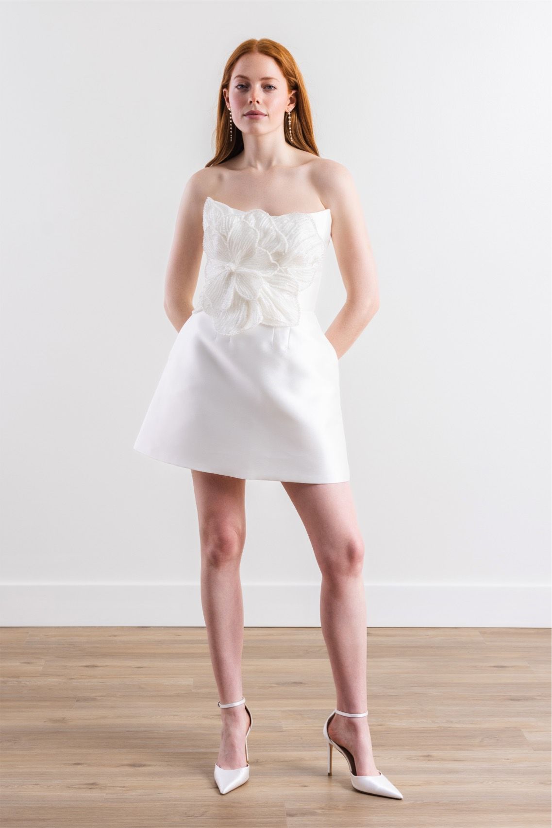 A woman in a white dress and heels is standing on a wooden floor.
