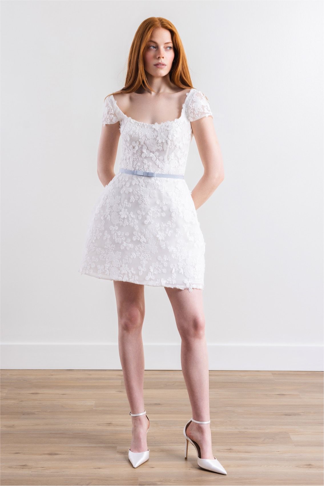 A woman is wearing a short white lace dress and heels.