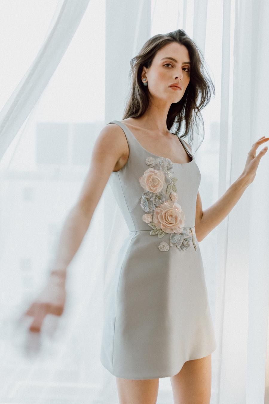 A woman in a grey dress is standing in front of a window.