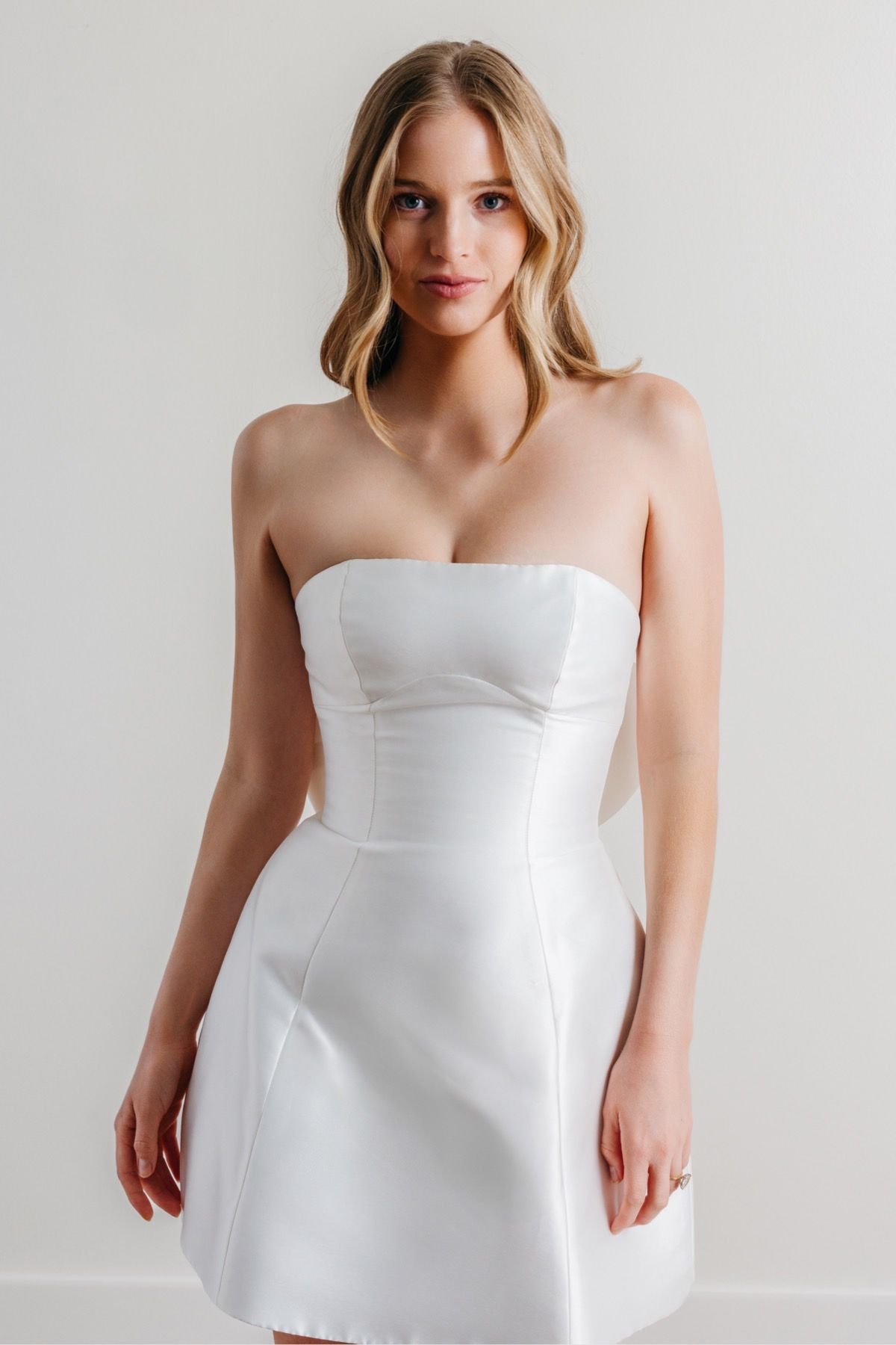 A woman in a white strapless dress is standing in front of a white wall.