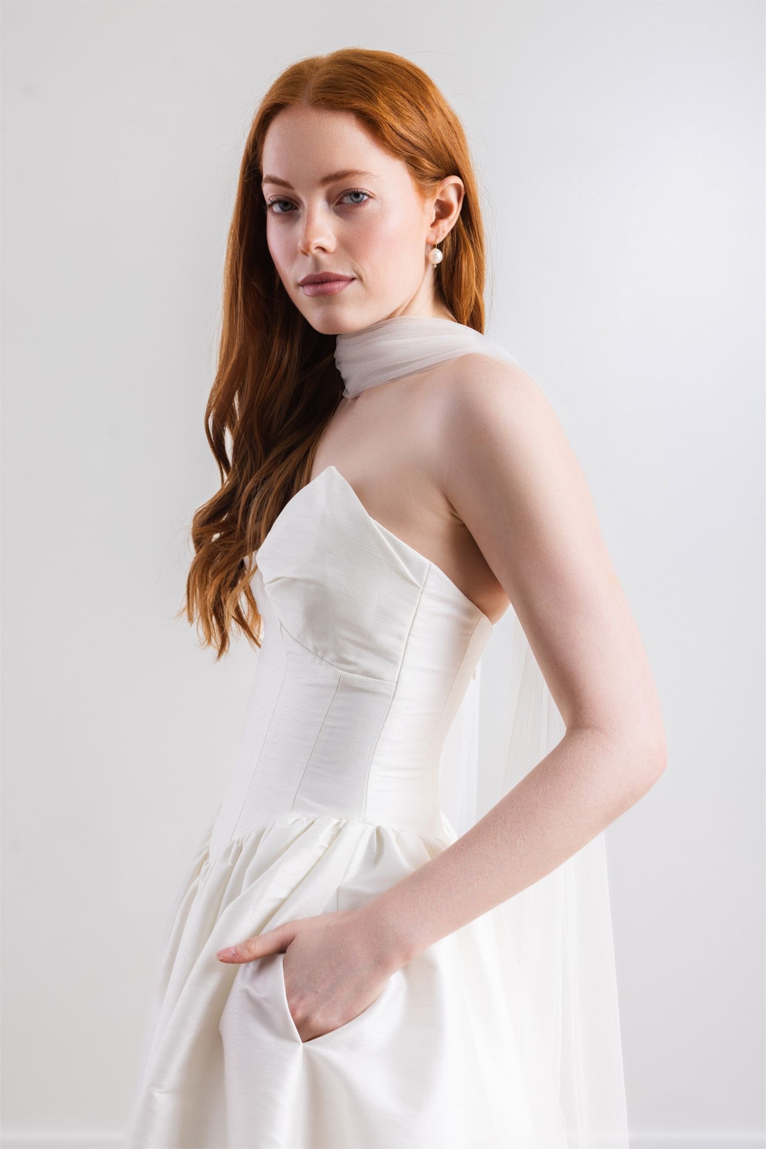 A woman in a white wedding dress is standing with her hands in her pockets.