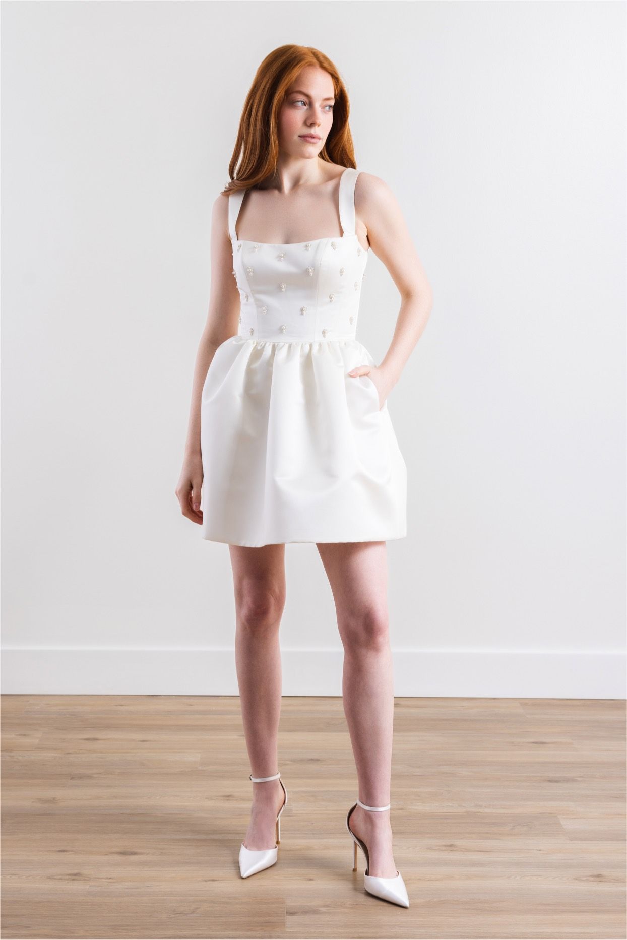 A woman in a white dress and heels is standing on a wooden floor.