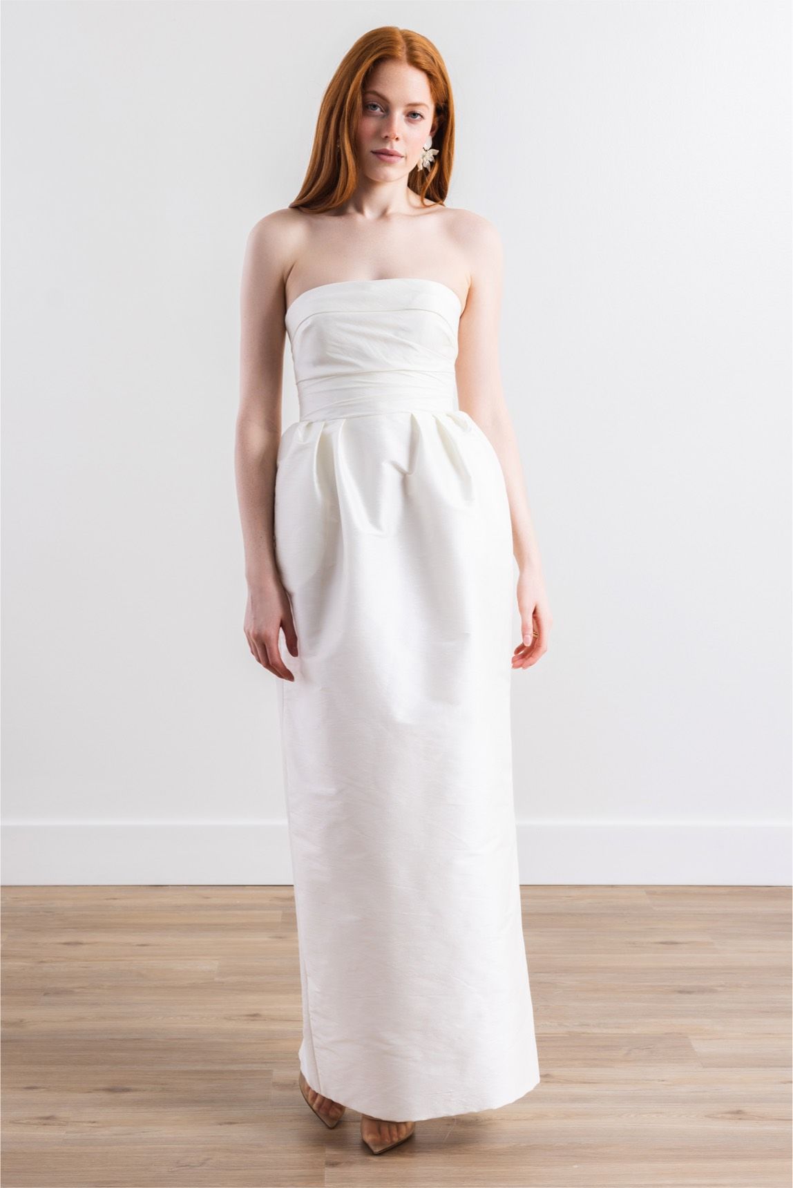 A woman in a white strapless dress is standing on a wooden floor.