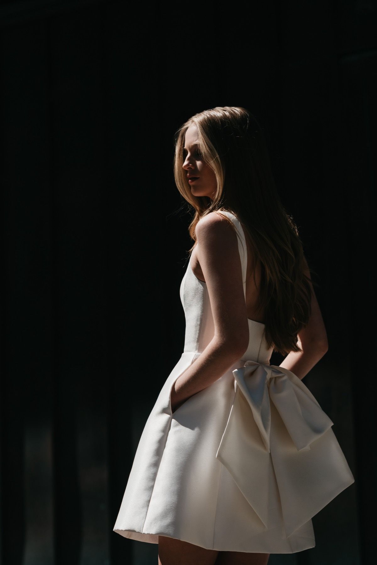 A woman in a white dress with a bow on the back is standing in the dark.
