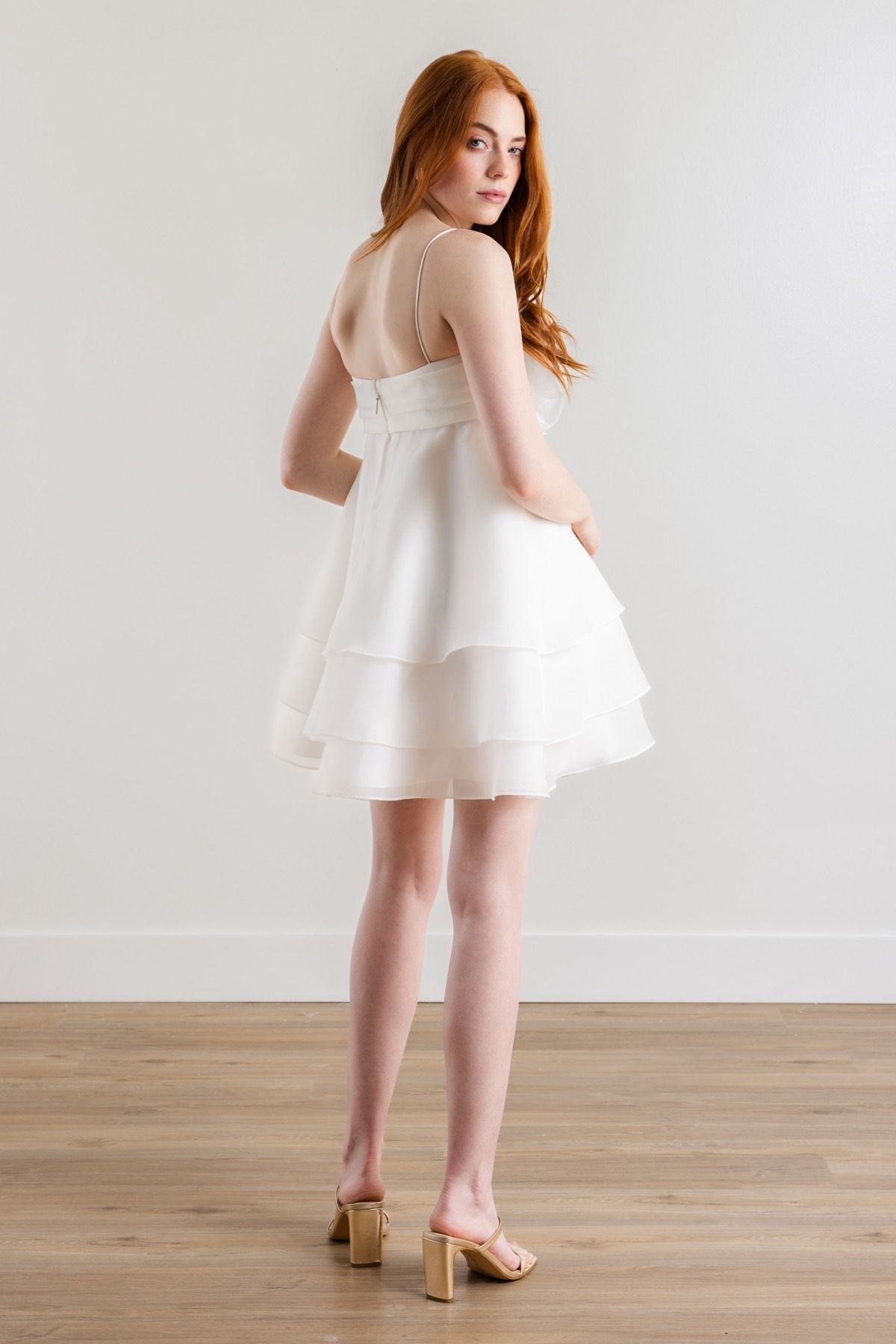 A woman in a white dress and heels is standing on a wooden floor.
