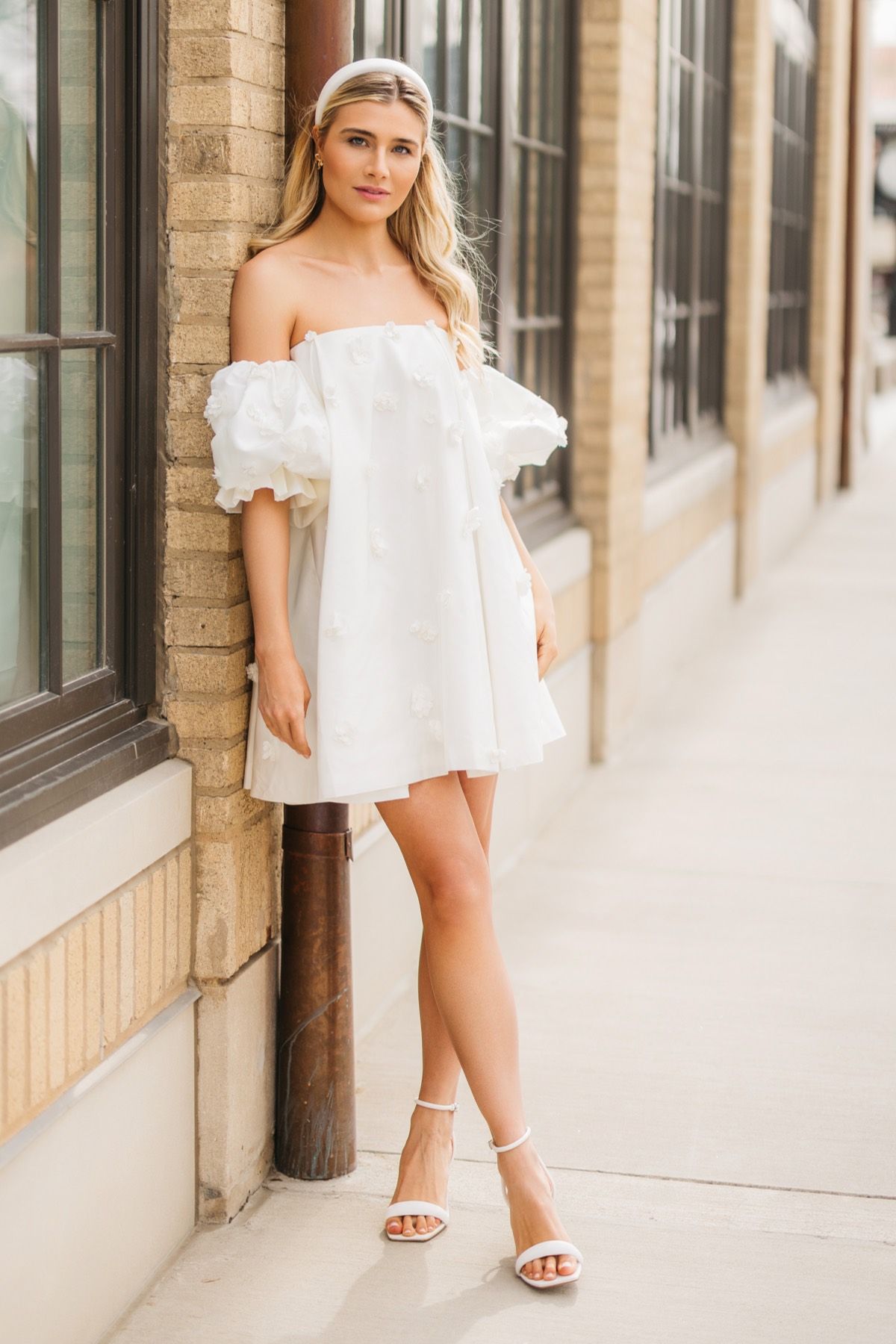 A woman in a white off the shoulder dress is standing in front of a building.