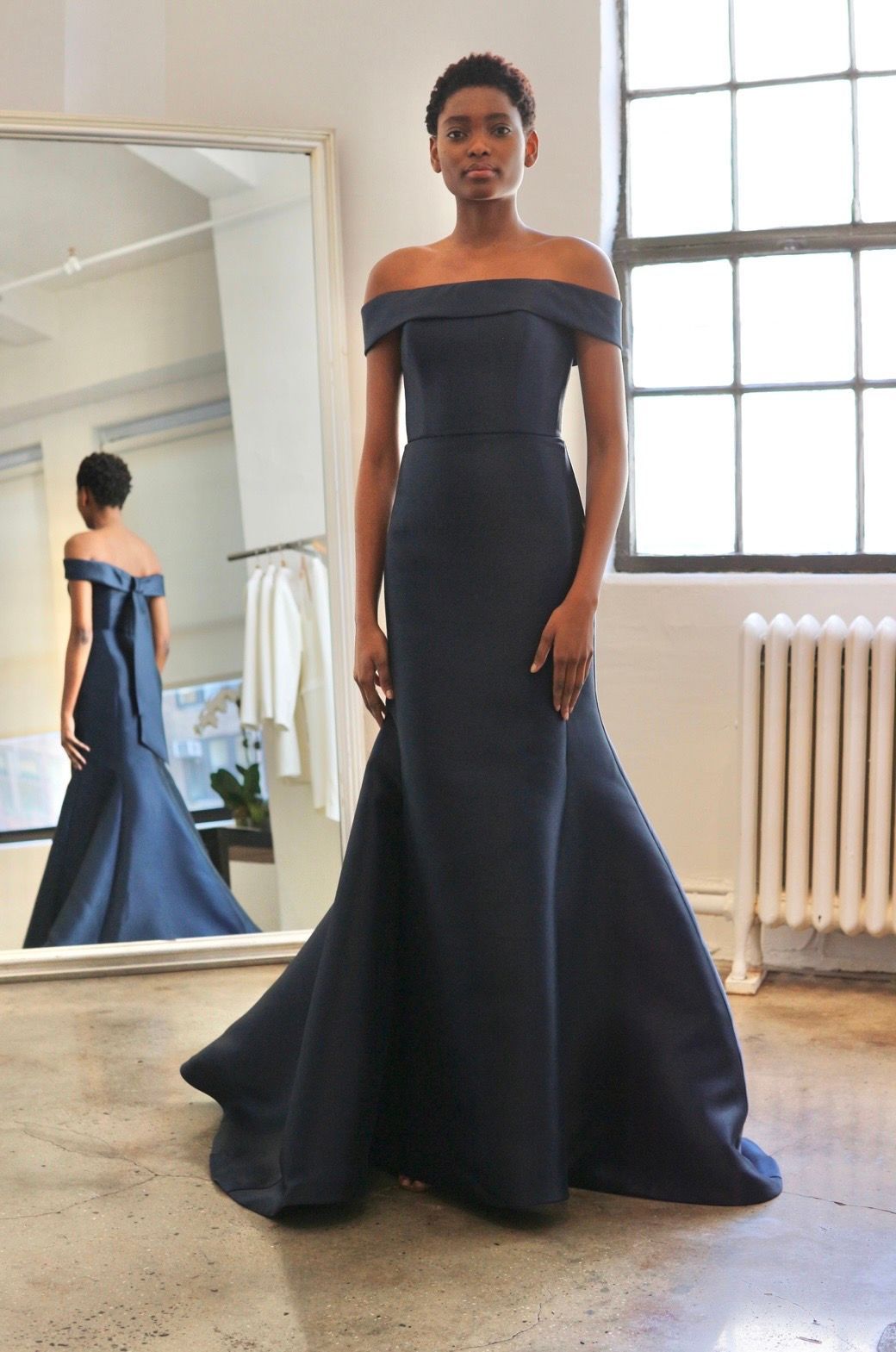 A woman in a blue off the shoulder dress is standing in front of a mirror.